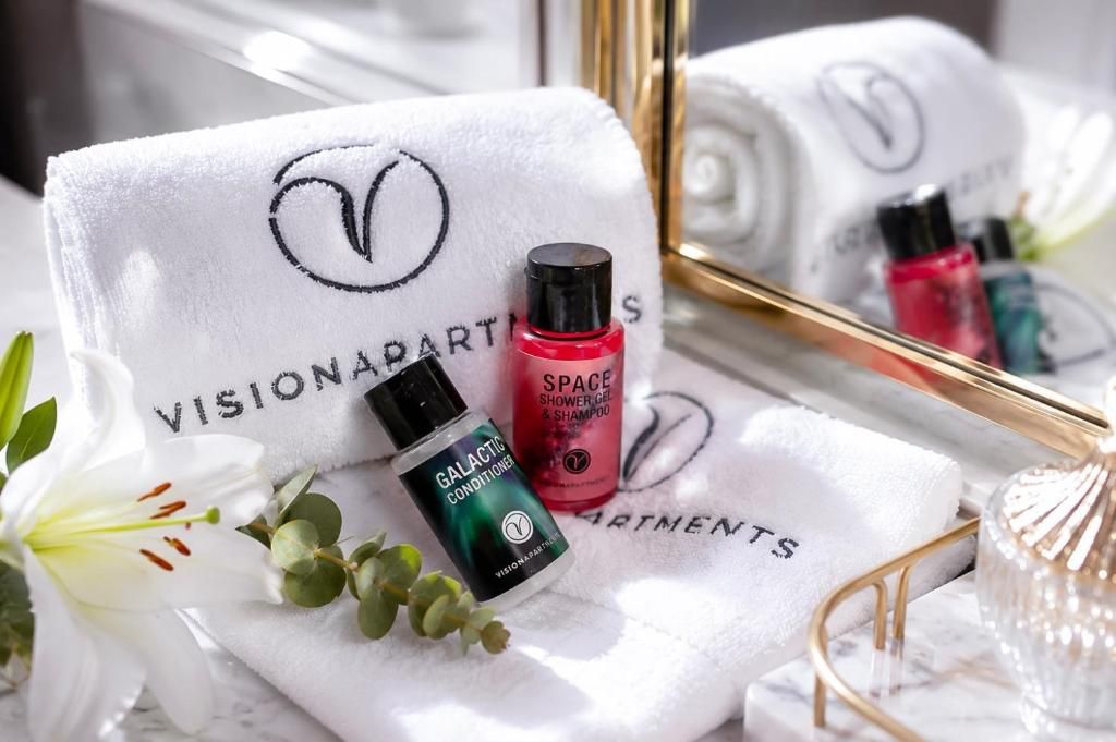 A towel with 'VISIONAPARTMENTS' branding, a 'SPACE SHOWER GEL & SHAMPOO' bottle, and a 'GALACTIC CONDITIONER' bottle.