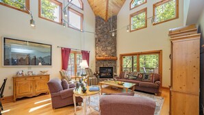 A spacious living room with a stone fireplace, wooden furniture, and large windows offering views of the outdoors. - Rustic-Meets-Modern Cabin Near Deep Creek (McHenry)