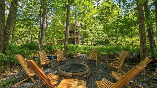 A forested area with a circular fire pit surrounded by wooden chairs and a wooden deck. - Rustic-Meets-Modern Cabin Near Deep Creek (McHenry)