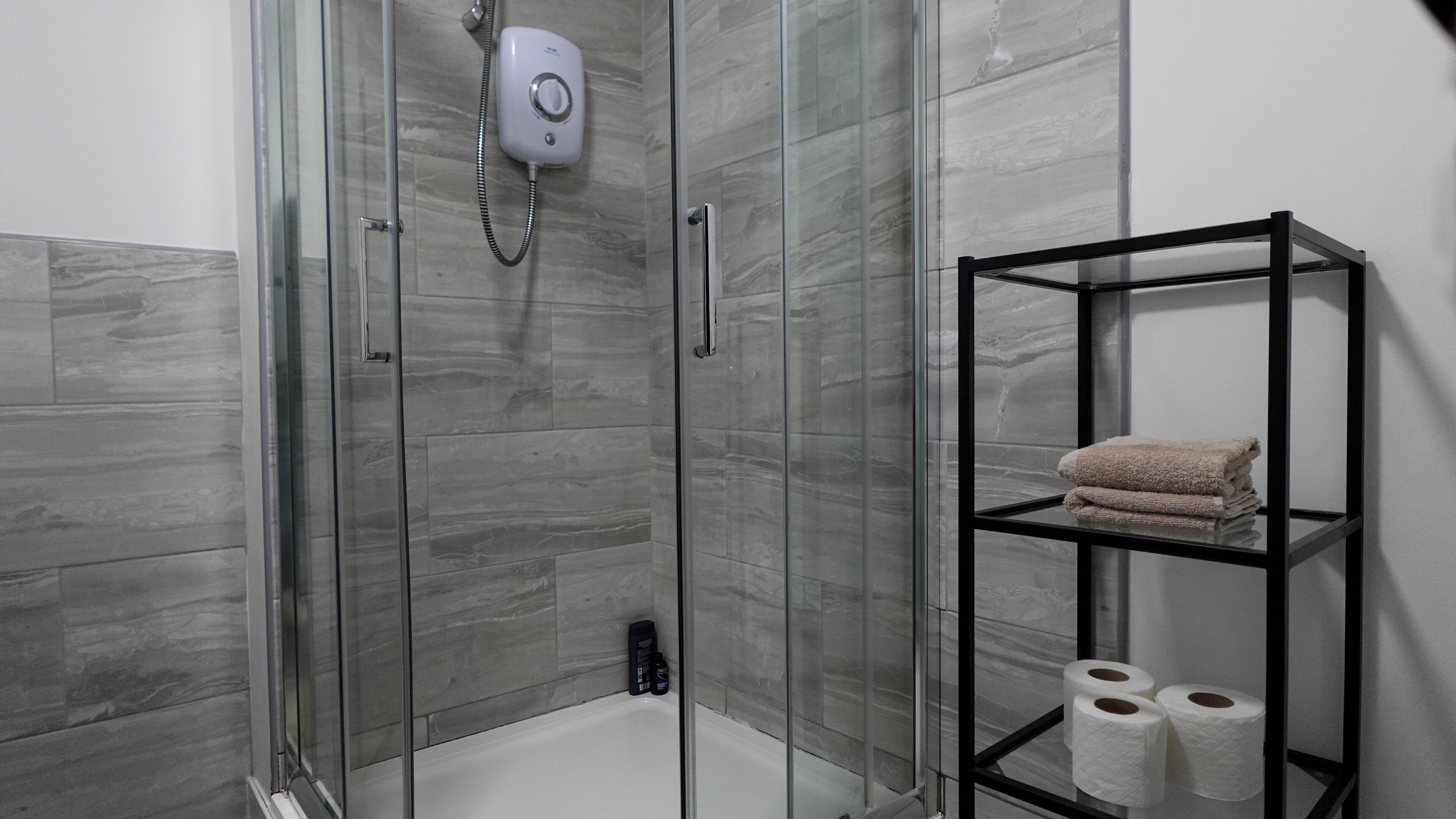Une salle de bain moderne avec une douche à parois de verre, une étagère en métal noir où sont posées des serviettes et un rouleau de papier toilette.