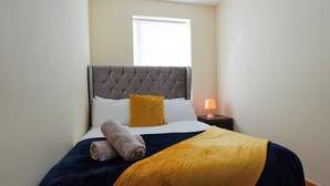 A neatly made bed with a grey upholstered headboard, a yellow throw blanket, and a rolled towel. There is a nightstand with a lamp and a window with blinds in the background. - The Stay Luxe (Manchester)