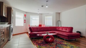 A modern living room with a red leather sofa, a small coffee table, and a colorful rug. There is a kitchen area with a washing machine and cabinets. - The Stay Luxe (Manchester)