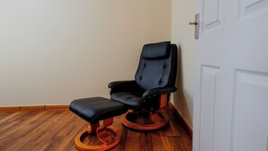A black massage chair with wooden base and orange accents, positioned in a corner of a room with wooden flooring and a white door. - The Stay Luxe (Manchester)