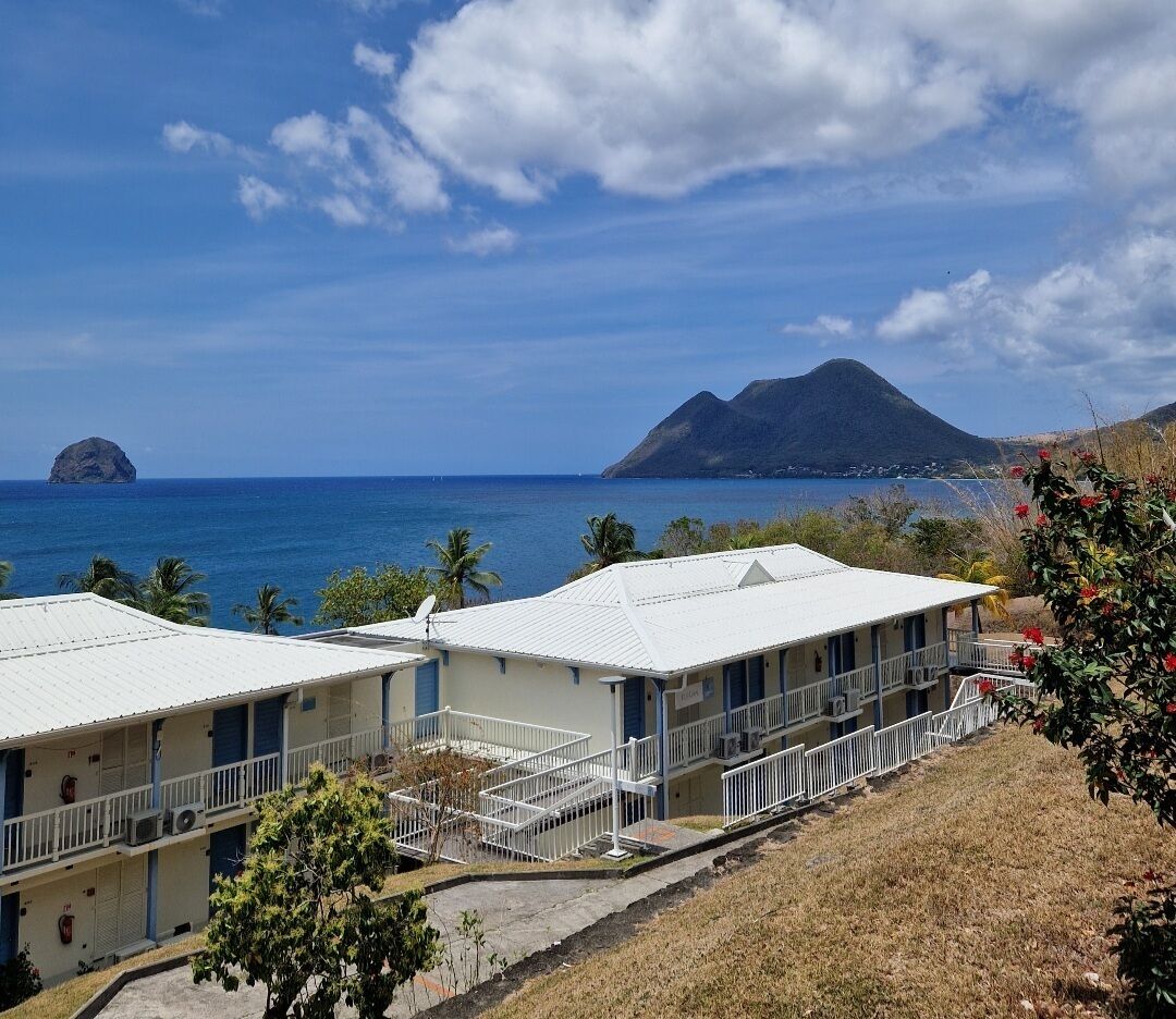 A coastal hotel with two-story buildings, a balcony, and a view of the sea and a mountain.