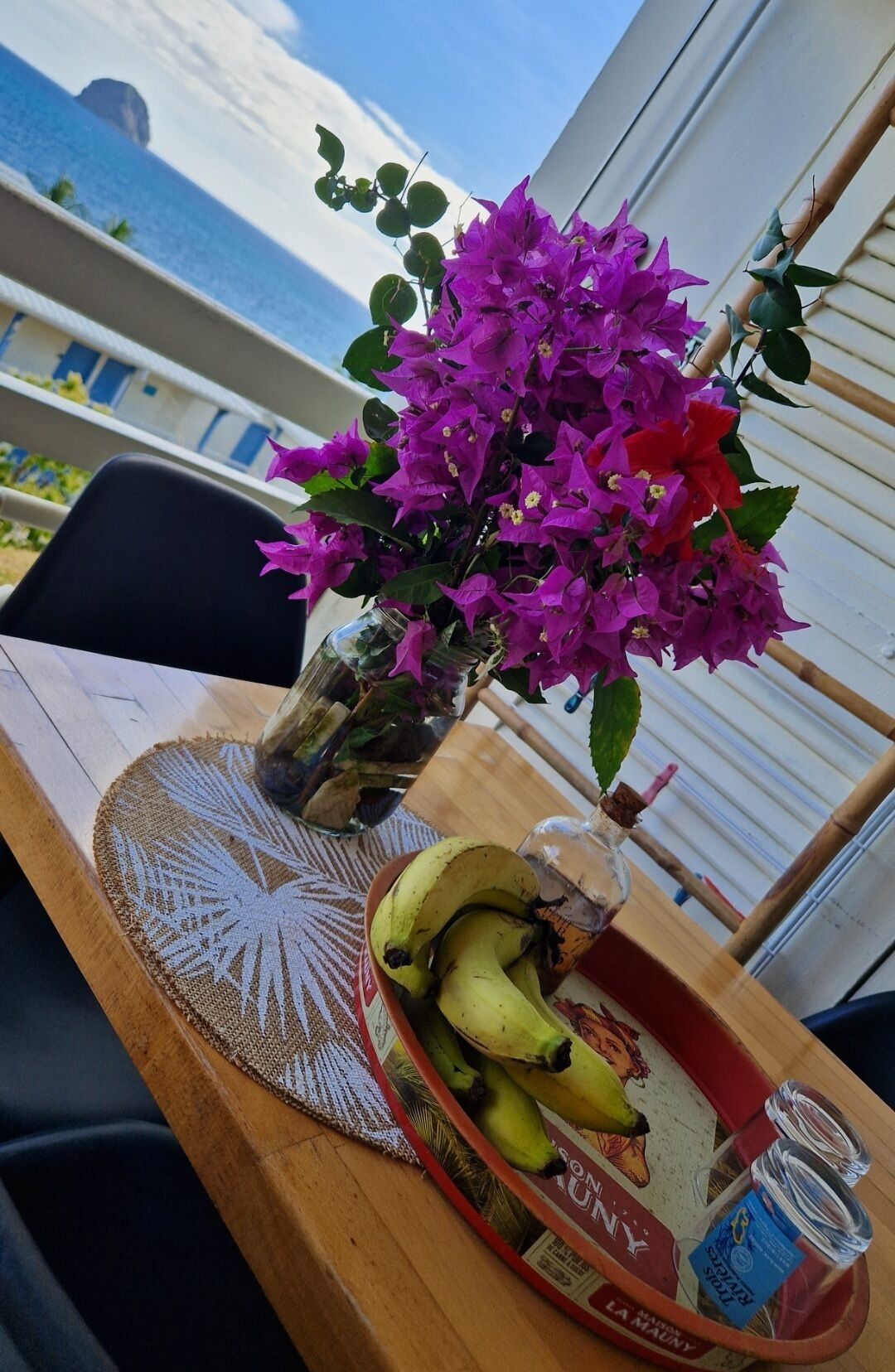 Une table ornée d’un vase de fleurs violettes, d’une assiette de bananes et d’une bouteille d’eau, le tout placé sur un balcon offrant une vue sur la mer.