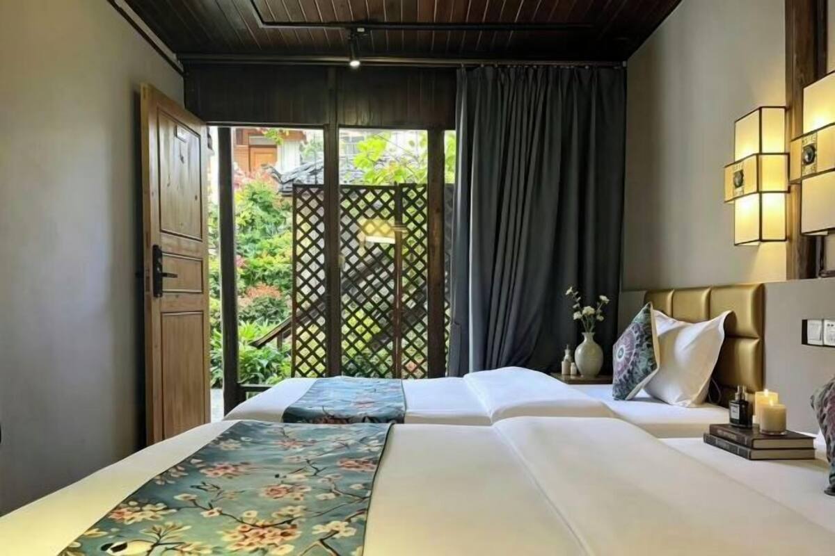 A hotel room with two beds, a view of greenery through a lattice door, and wall-mounted lighting.