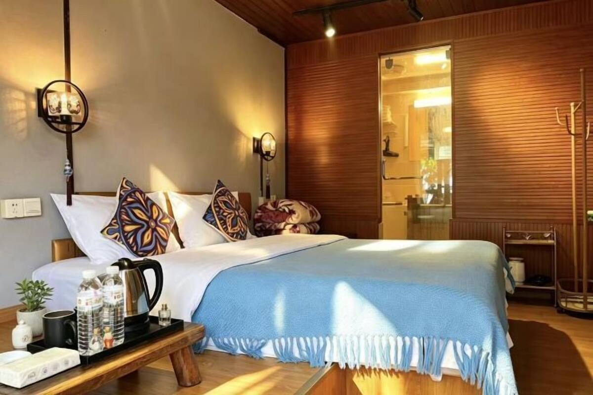 A neatly made bed with a blue bedspread, a bedside table with a kettle and water bottles, and a wooden wall with a sliding door.