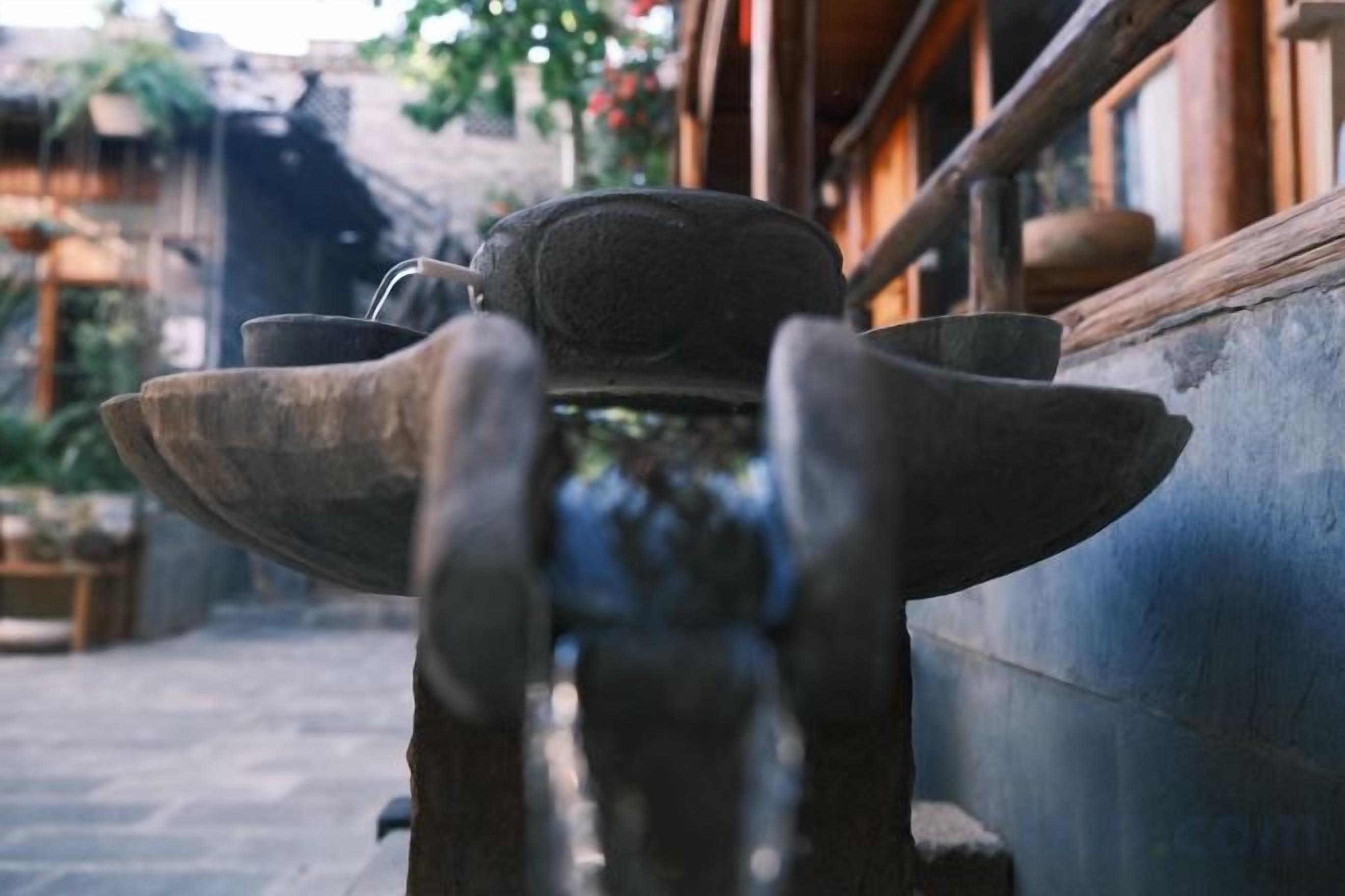 A traditional water fountain with a stone basin and a metal spout, located in an outdoor courtyard with a tiled floor and wooden furniture.