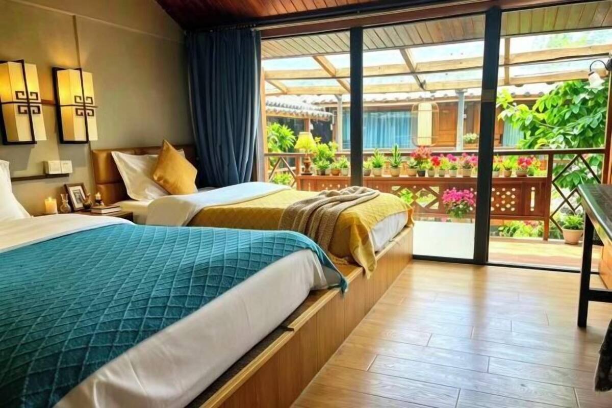 A hotel room with two beds, a wooden floor, a balcony with potted plants, and a view of the garden.