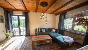 A cozy living room with a wooden ceiling, a green sofa, a coffee table, and a large window with a view of greenery. - Gemütliche Ferienwohnung in der Vulkaneifel Casa Oos (Rockeskyll)