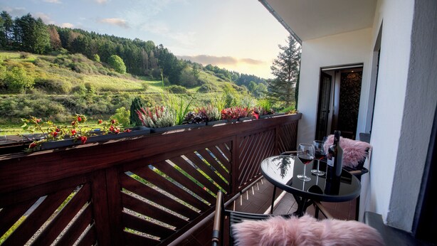 A balcony with a view of a green hillside, a table with wine glasses, and a chair. - Gemütliche Ferienwohnung in der Vulkaneifel Casa Oos (Rockeskyll)