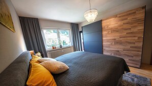 A bedroom with a large bed, grey and yellow pillows, a window with curtains, a wooden wall panel, and a chandelier. - Gemütliche Ferienwohnung in der Vulkaneifel Casa Oos (Rockeskyll)
