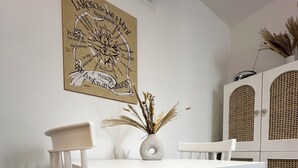 Un área de comedor con una mesa y sillas blancas, un jarrón decorativo con plantas secas y una obra de arte colgada en la pared.