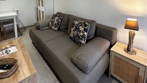 Una sala de estar moderna con un sofá gris, dos almohadones estampados, una mesita de madera con una lámpara y un mueble blanco decorado con objetos.