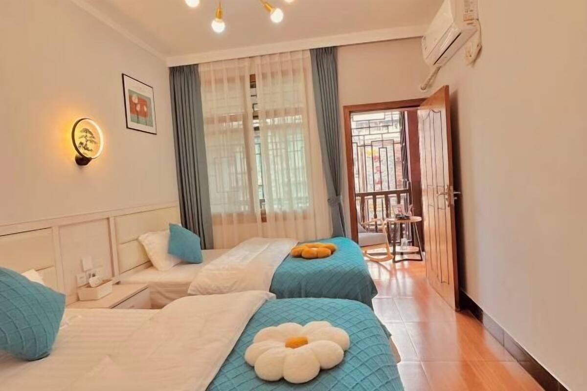 A hotel room with two beds, a small table, a wall-mounted light, and a balcony with a railing.