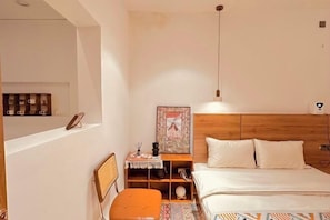 A bedroom with a wooden headboard, a bed with white bedding, a small table with a lamp, and a shelf with decorative items.