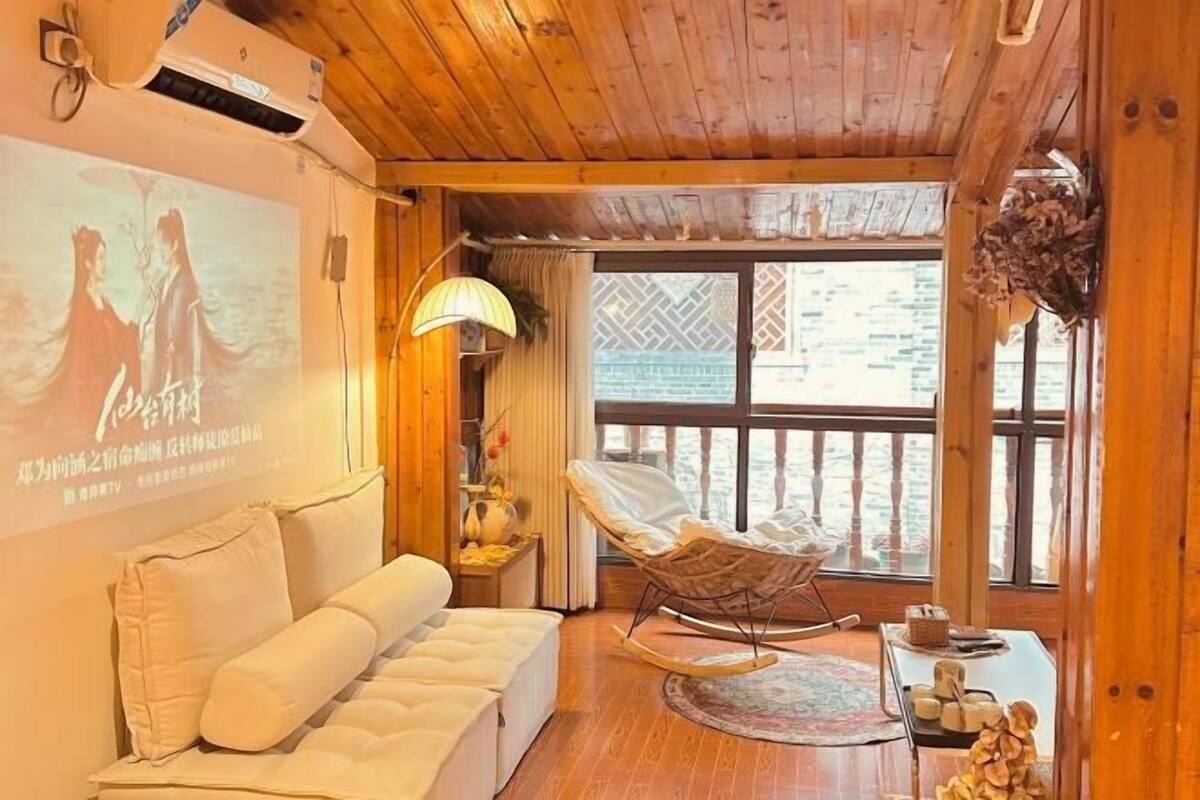 A cozy room with a wooden ceiling, a white sofa, a rocking chair, and a large window with a view.