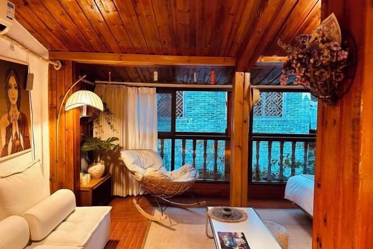 A cozy room with wooden walls, a white sofa, a rocking chair, a small table, and a view through a window with a brick wall.