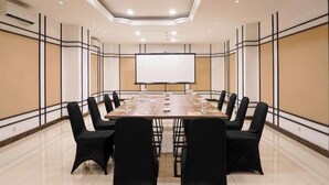 Een vergaderzaal met een lange tafel, zwarte stoelen, een projectiescherm en een betegelde vloer.