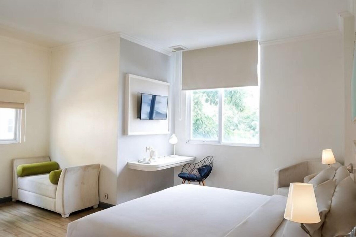 Ett hotellrum med en säng, två stolar, ett skrivbord, en tv och ett fönster.
