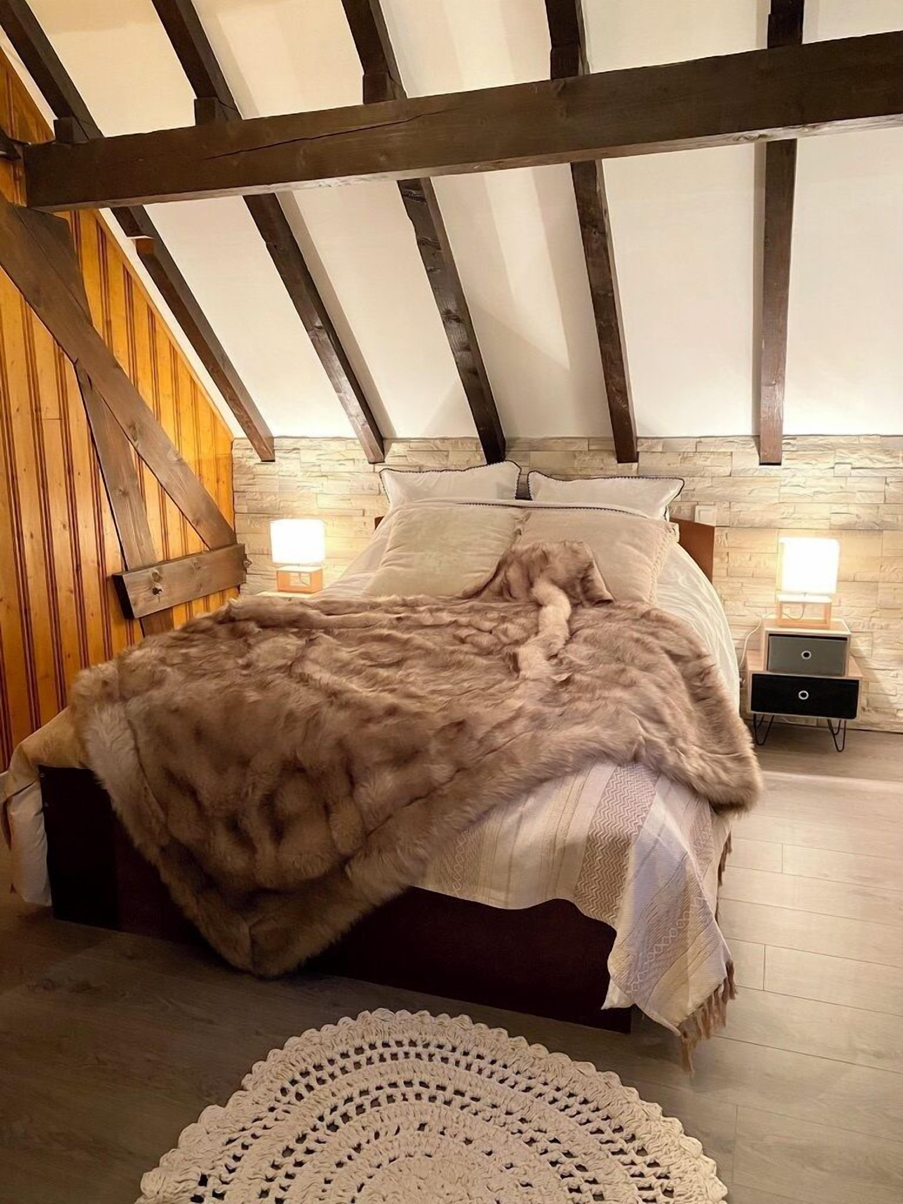 Un dormitorio con techo de madera, pared de piedra, cama con manta de piel y lámparas de noche.