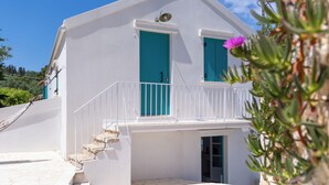 Un bâtiment blanc avec des portes et des fenêtres de couleur turquoise, un balcon doté de balustrades blanches et un escalier en pierre menant à l’entrée.