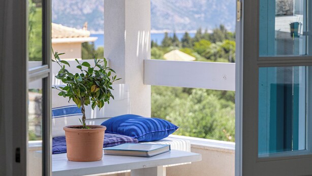 Une porte ouverte donnant sur un balcon où se trouvent un pot de fleurs, un coussin bleu et une vue sur les montagnes.