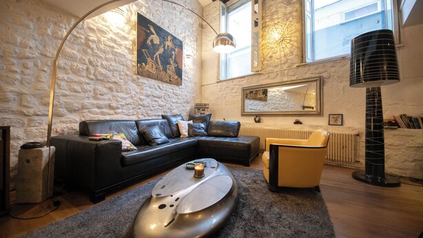 A cozy living room with a stone wall, a black leather sofa, a modern floor lamp, and a unique metallic sculpture. - Your stay in the heart of Paris in a large duplex apartment. Saint Germain (Paris)