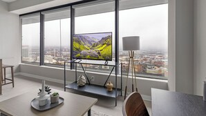 A modern living room with a large window, a flat-screen TV on a stand, a dining area with a table and chairs, and a floor lamp.