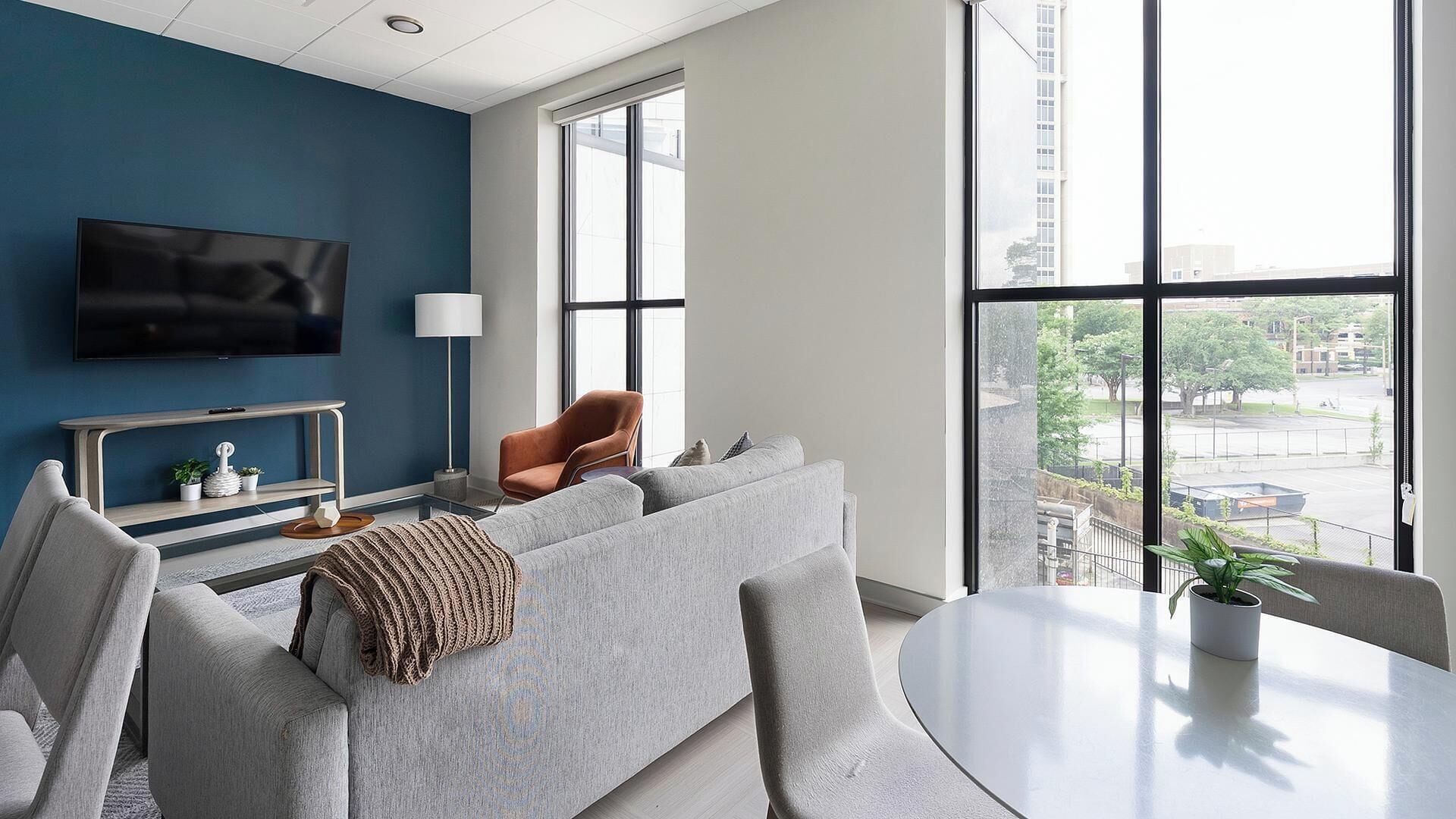 Un salon moderne avec un canapé gris, un fauteuil marron, un téléviseur et une grande fenêtre donnant sur la ville.