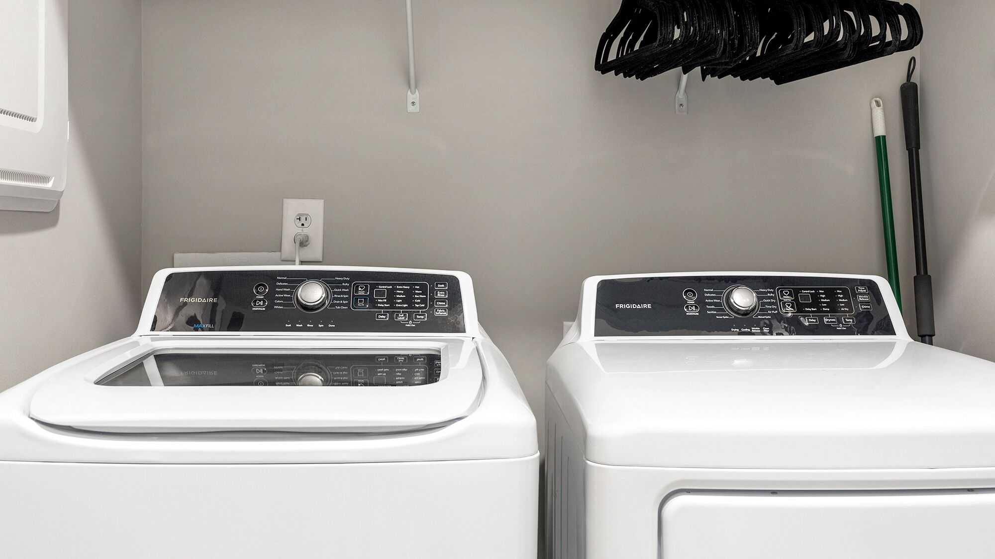 Two white front-loading washing machines with control panels, a power outlet, and a green mop on the wall.