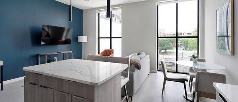 A modern kitchen with a marble countertop, wooden cabinets, a built-in TV, and a large window offering a view of the outdoors.