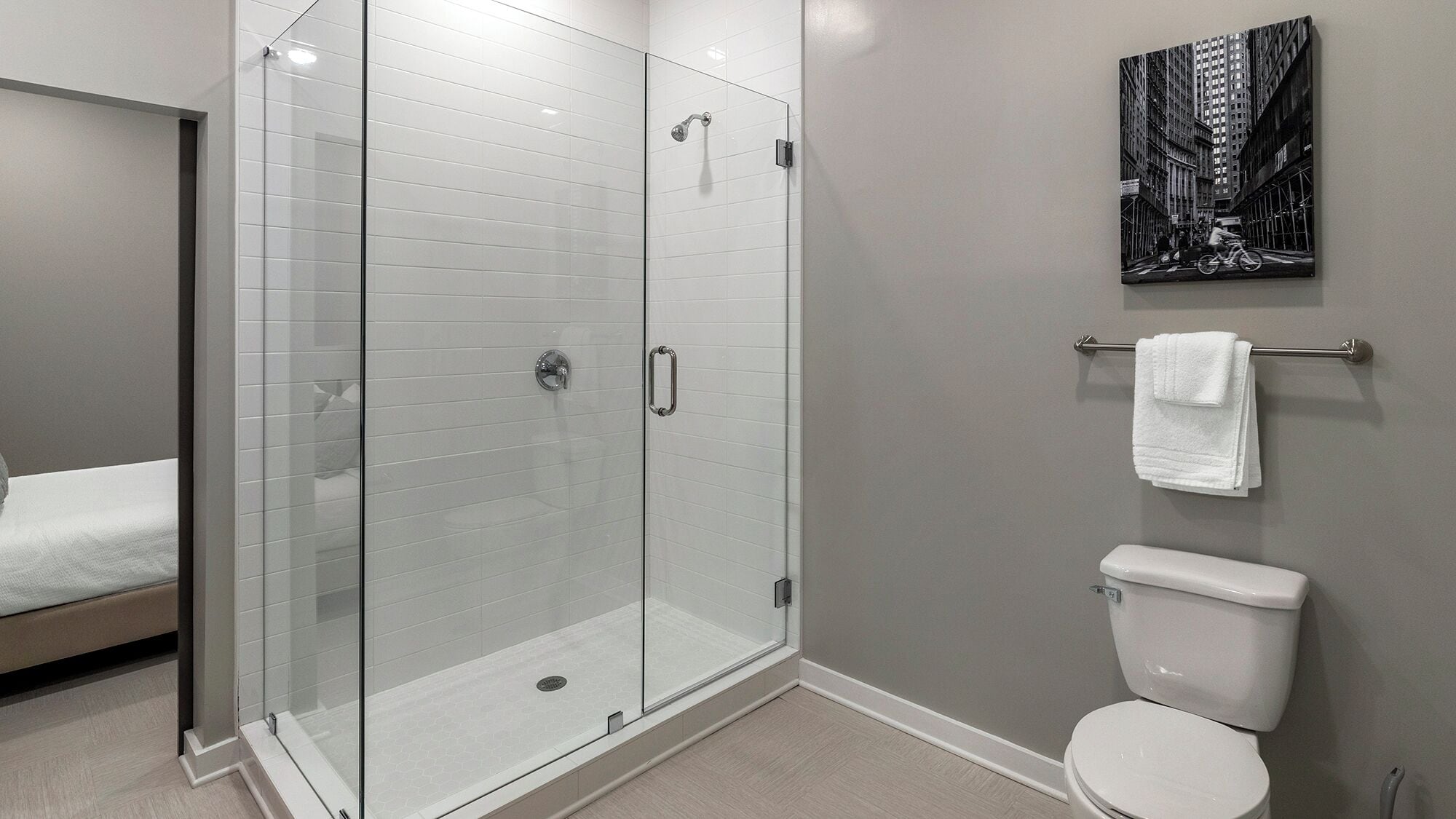 A modern bathroom with a glass-enclosed shower, a toilet, and a bed in the adjacent room.