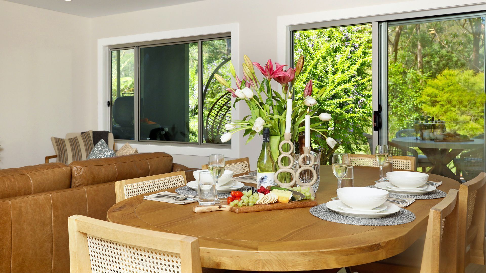 A dining area with a round wooden table set for four, surrounded by a brown leather sofa and wooden chairs. A vase with flowers and a candle is on the table.