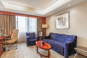 Ein Hotelzimmer mit einer Couch, zwei Sesseln, einem kleinen Tisch, einem Schreibtisch mit Stuhl, einem Fernseher und einem gerahmten Bild an der Wand.