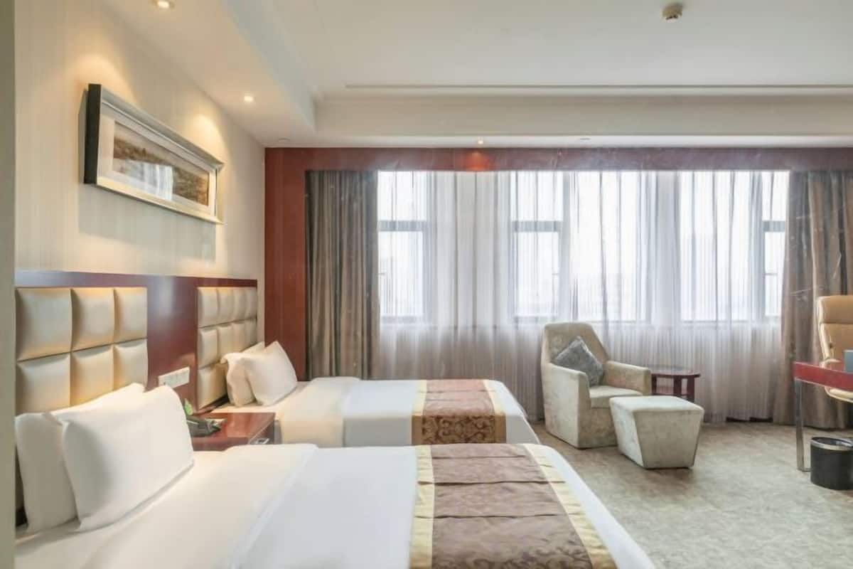 A hotel room with two beds, a sitting area with a chair and a small table, a large window with curtains, and a framed picture on the wall.