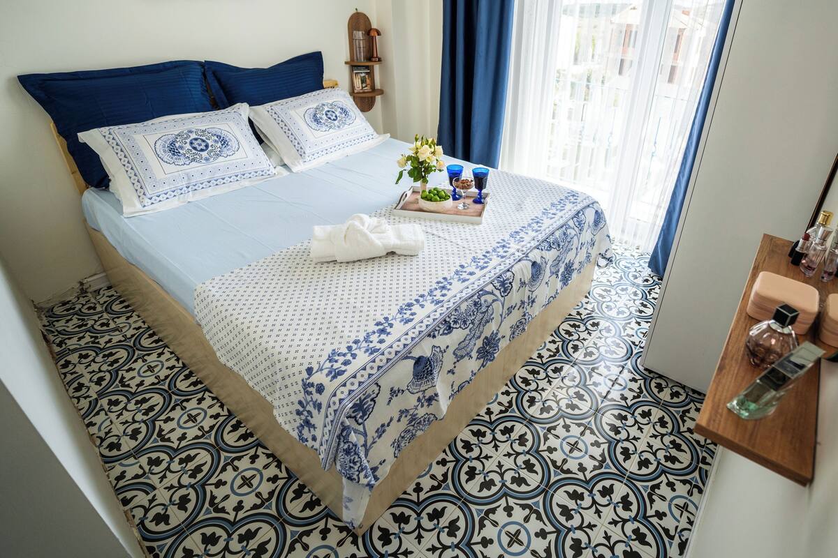 A neatly made bed with blue and white bedding, a small bedside table with toiletries, and patterned floor tiles.