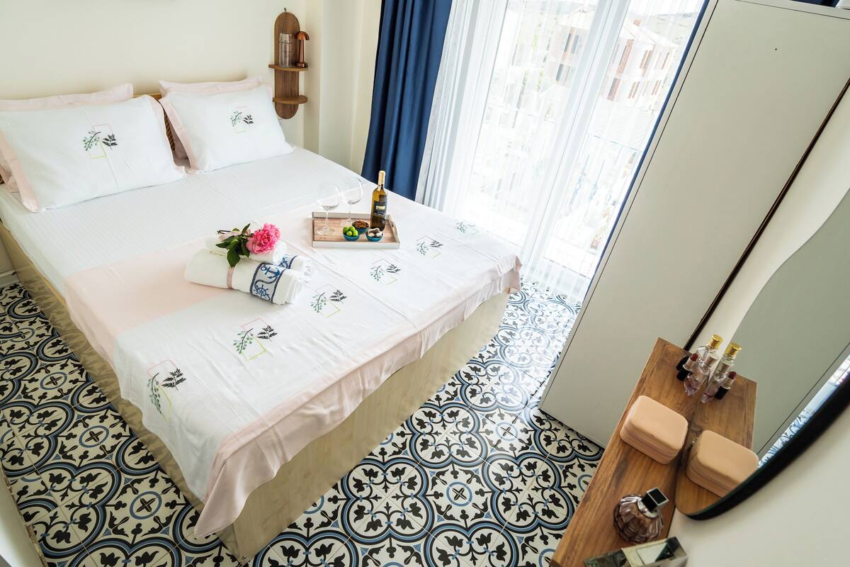 A neatly made bed with a white bedspread and matching pillows, a tray with a bottle, glasses, and a flower arrangement on the bed.