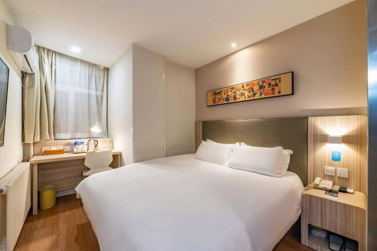 A hotel room with a large bed, a desk with a chair, a small table with a lamp, a wall-mounted air conditioner, and a painting above the bed.