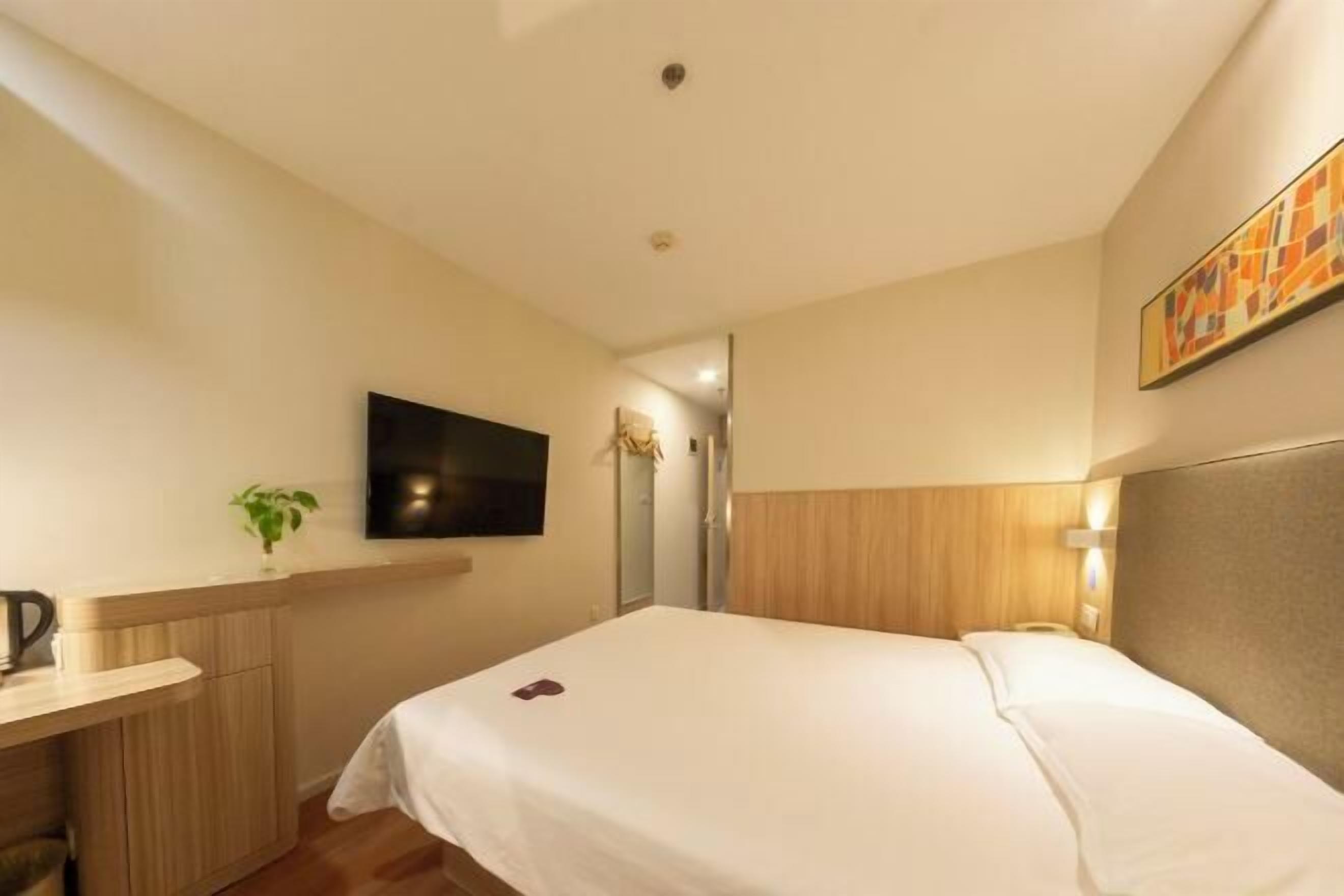 A hotel room with a bed, a desk with a plant, a flat-screen TV, and a painting on the wall.