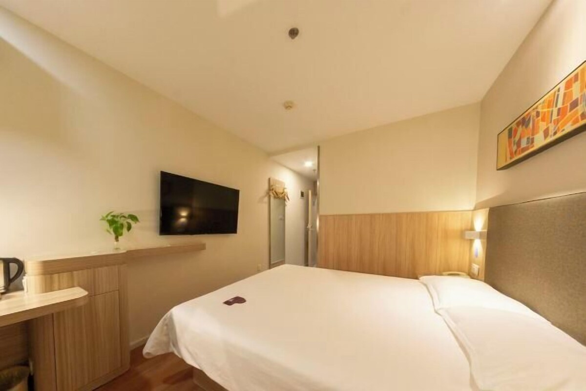 A hotel room with a bed, a desk with a plant, a flat-screen TV, and a painting on the wall.