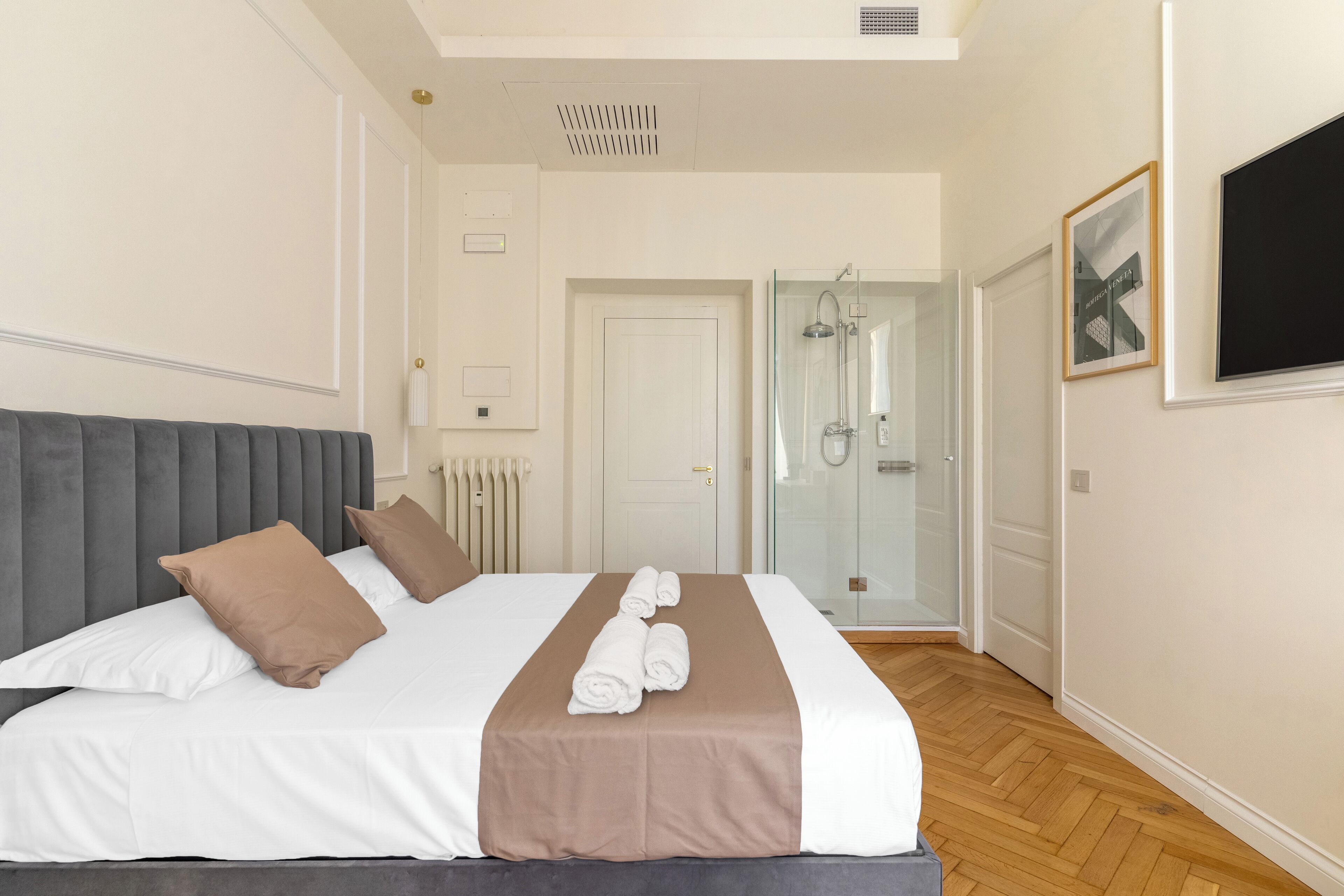 A neatly made bed with a grey headboard, white and brown pillows, and rolled towels.