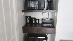 Una kitchenette compacta con un refrigerador negro, un microondas, una cafetera y un tostador sobre una estantería de madera.