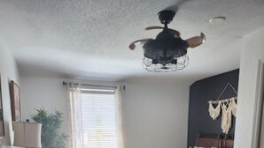 Un dormitorio pequeño con cama, ventilador de techo, mesita de noche, lámpara, ventana con cortinas y una planta pequeña.