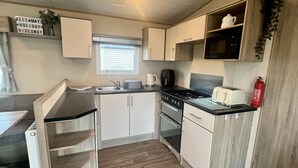 A compact kitchen with a stove, sink, and a toaster. There is a window with blinds, a shelf with a plant, and a sign on the wall. - Immaculate 3-bed Static Caravan in Skegness (Skegness)