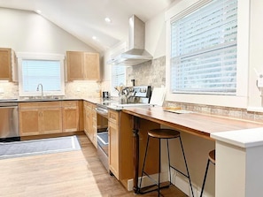 A kitchen with wooden cabinets, a stainless steel range, a window with blinds, and a bar stool. - The Almost There | Conway NH (Conway)