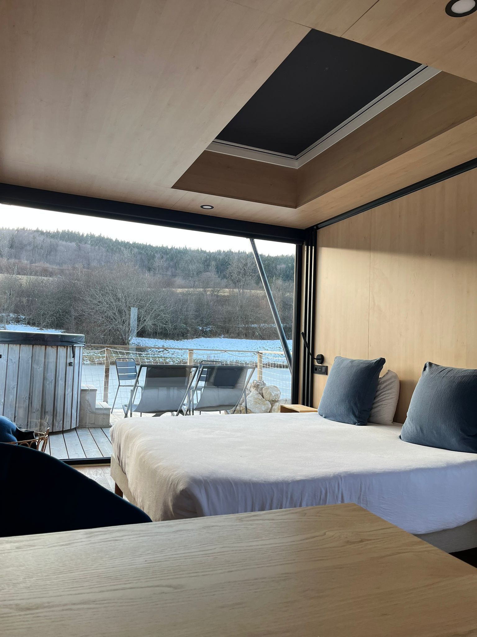 Habitación moderna con una cama, dos almohadas, una mesa de madera y una bañera de hidromasaje visible a través de la ventana.