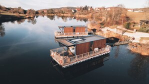 A floating houseboat with a deck and a covered area, situated on a calm lake with a backdrop of trees and houses. - Bleu Minuit - La Rive Etoilée (Plateau d'Hauteville)