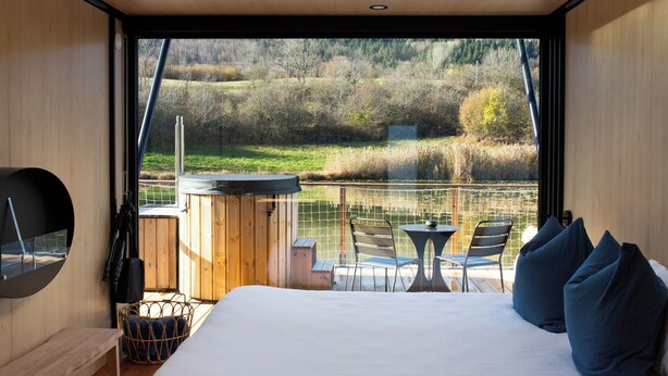 A bedroom with a large bed, a wooden desk, a chair, and a small table with a chair. There is a window with a view of a lake and trees. - Bleu Minuit - La Rive Etoilée (Plateau d'Hauteville)