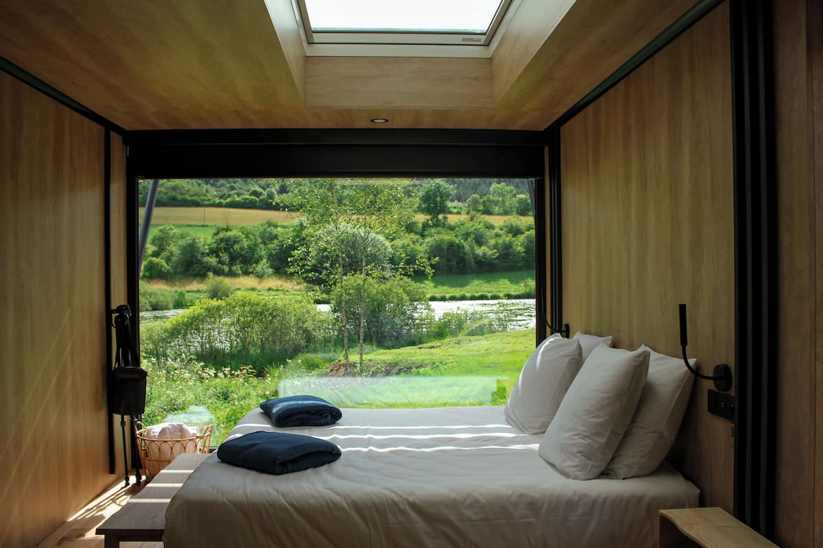 A bedroom with a large window, a bed with pillows, a bedside table, and a view of a green landscape.